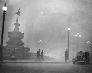 December 1952: London during the day during the Great Smog event. A black-and-white photo of a London street. The air is hazy, filled with smog. People walk along the sidewalk beside a fountain, and an old-fashioned car is parked by the curb. Streetlights line the sidewalk, with some in the distance only faintly visible. Buildings in the background are almost invisible through the smog.