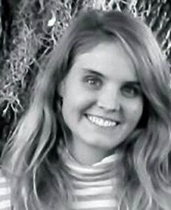 Isobel Pribil's headshot, in black and white. She is a young blonde woman wearing a striped turtleneck, standing outdoors. She is smiling at the camera.