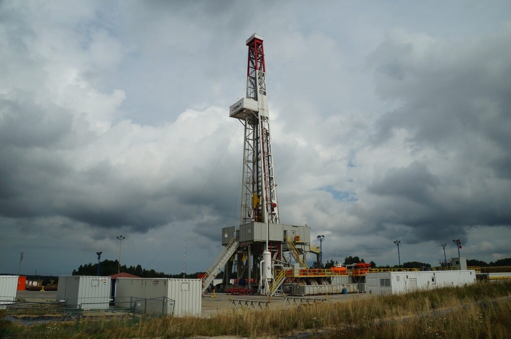 A red-and-white drill tower stands surounded by trailers and equipment, against a cloudy sky.