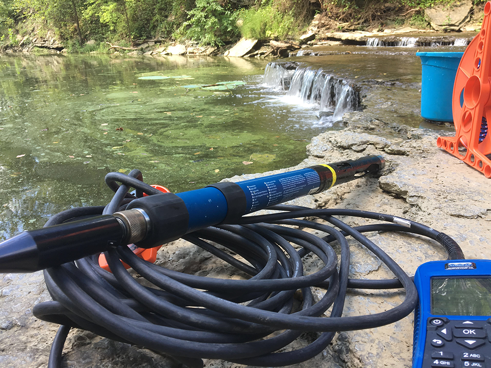 A tool resting on a coiled hose sits beside a river with algae in it. A small waterfall is in the background, along with other tools like a blue bucket.